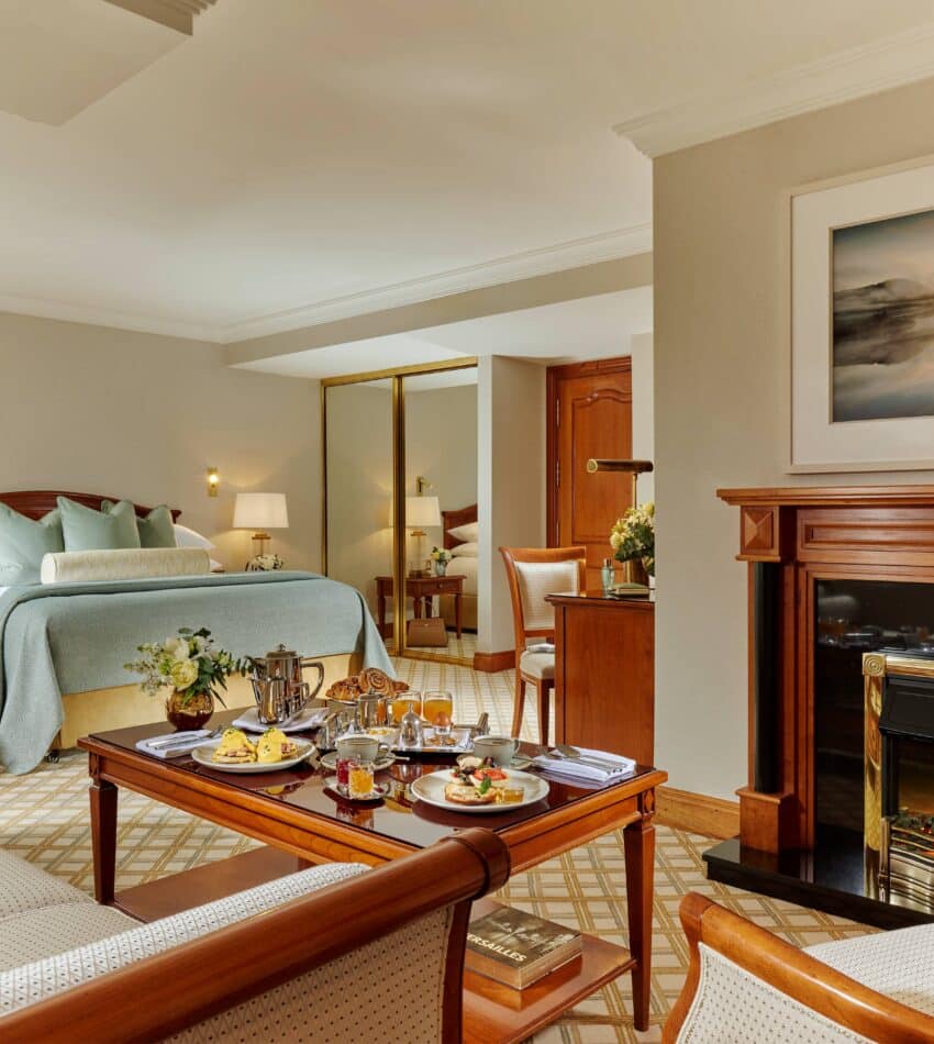 A hotel suite with a bed, seating area, and coffee table set for breakfast. There is a fireplace with a landscape painting above it and a large mirror on the wall.