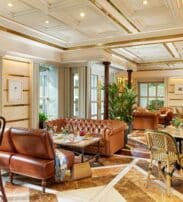 Elegant lounge area with leather sofas, armchairs, marble floor, indoor plants, and large windows letting in natural light.