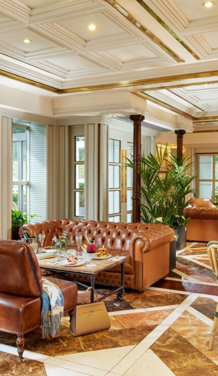 Elegant lounge area with leather sofas, armchairs, marble floor, indoor plants, and large windows letting in natural light.