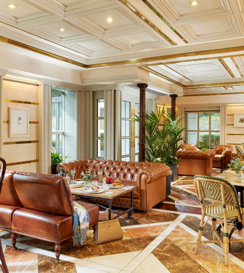 Elegant lounge area with leather sofas, armchairs, marble floor, indoor plants, and large windows letting in natural light.