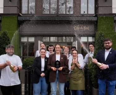 A group of people poses outside Café du Parc, with two men spraying champagne and a woman in the centre holding an award.