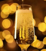 A close-up of champagne being poured into a flute glass, with golden bubbles rising and blurred warm lights in the background.
