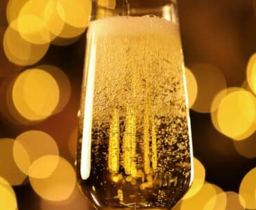 A close-up of champagne being poured into a flute glass, with golden bubbles rising and blurred warm lights in the background.