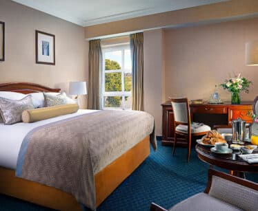 A hotel room with a large bed, desk, armchairs, and a window with curtains; breakfast items and flowers are set on a table near the bed.