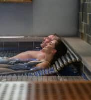 Two people relax in a swimming pool, leaning back against metal rails at the edge, with their heads above water and eyes closed.