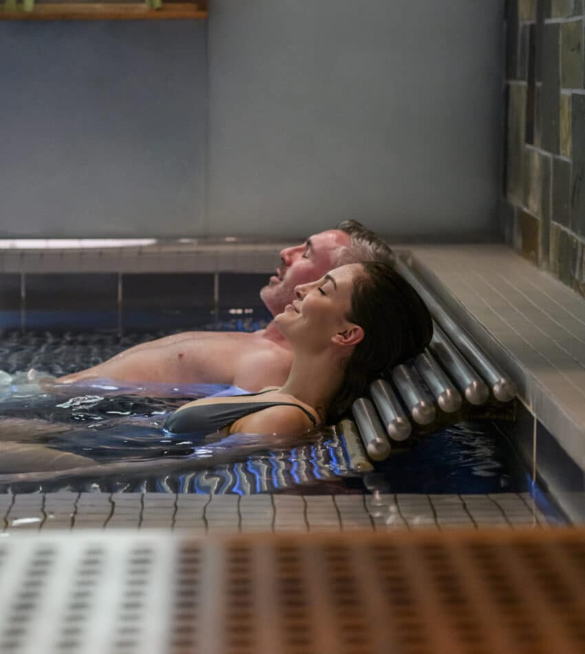 Two people relax in a swimming pool, leaning back against metal bars at the edge, with their heads resting and eyes closed.