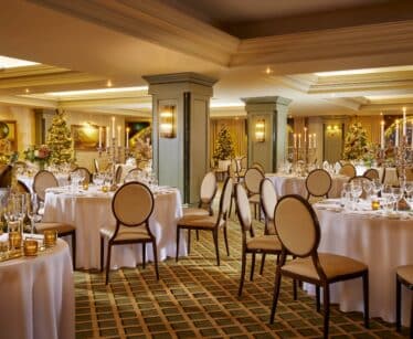 Elegant banqueting hall set for an event, with round tables covered in white cloths, arranged chairs, glassware, and festive decorations including lit Christmas trees.