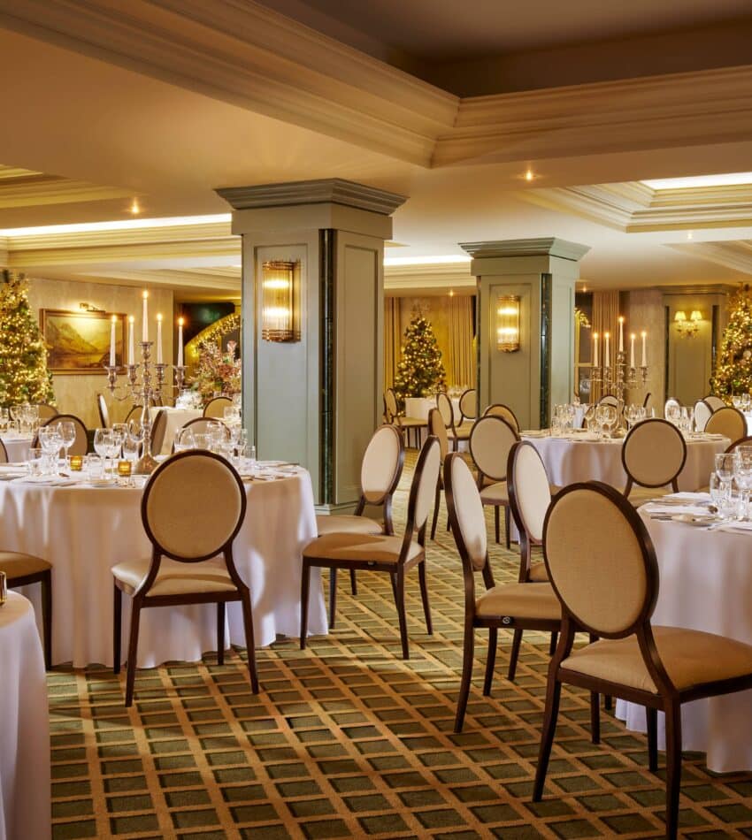 Elegant banqueting hall set for an event, with round tables covered in white cloths, arranged chairs, glassware, and festive decorations including lit Christmas trees.