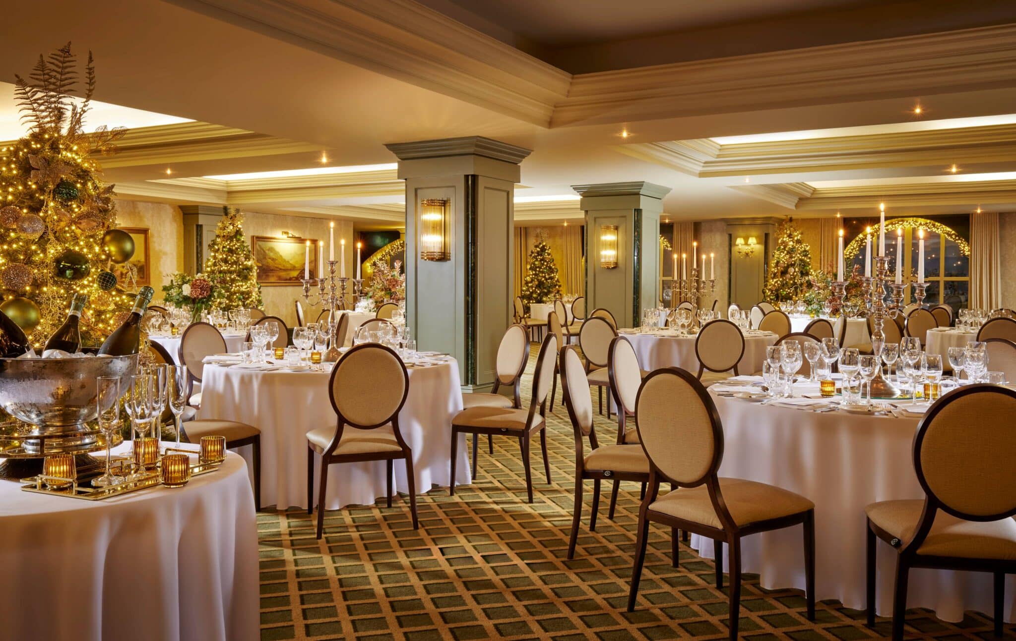 Elegant banqueting hall set for an event, with round tables covered in white cloths, arranged chairs, glassware, and festive decorations including lit Christmas trees.