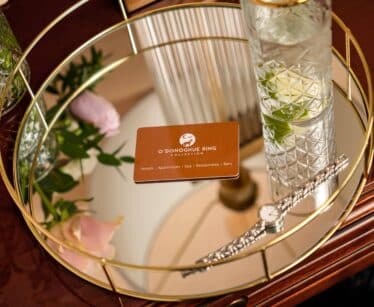 A round gold tray holds a drink with lime and mint, a silver watch, a pink flower, and an O'Donoghue Ring Collection card on a wooden surface.