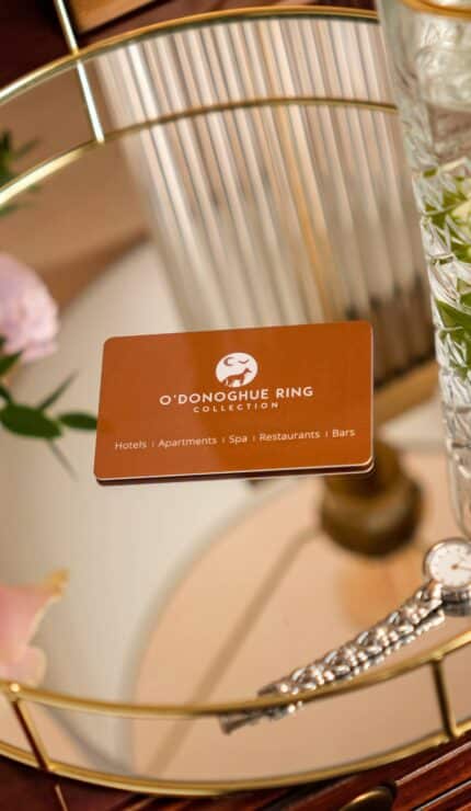 A round gold tray holds a drink with lime and mint, a silver watch, a pink flower, and an O'Donoghue Ring Collection card on a wooden surface.