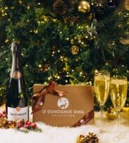 A bottle of Taittinger champagne, two filled glasses, and an O’Donoghue Ring Collection box are arranged on snow-like ground in front of a decorated Christmas tree.