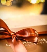 A close-up of a brown gift box tied with a shiny satin ribbon, with blurred warm lights in the background.