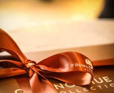 A close-up of a brown gift box tied with a shiny satin ribbon, with blurred warm lights in the background.