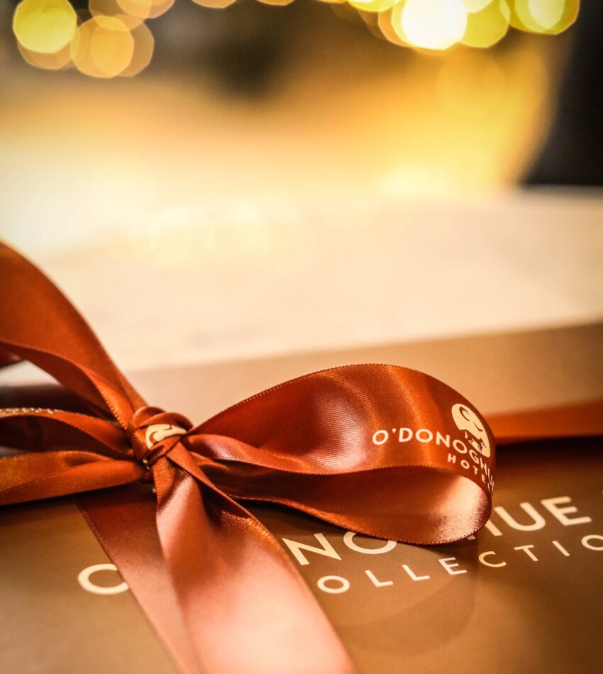 A close-up of a brown gift box tied with a shiny satin ribbon, with blurred warm lights in the background.