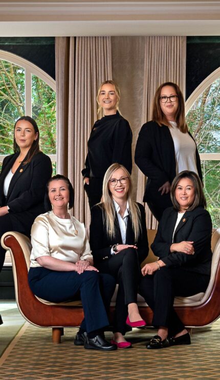 Group of women in business attire pose together in a well-lit, elegant room with large windows and armchairs.