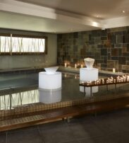Indoor spa with tiled walls and floor, a pool, three illuminated white sculptures, candles on a wooden platform, and soft overhead lighting.