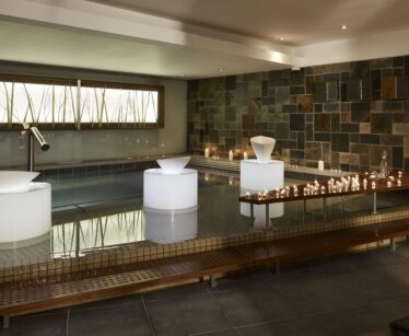 Indoor spa with tiled walls and floor, a pool, three illuminated white sculptures, candles on a wooden platform, and soft overhead lighting.