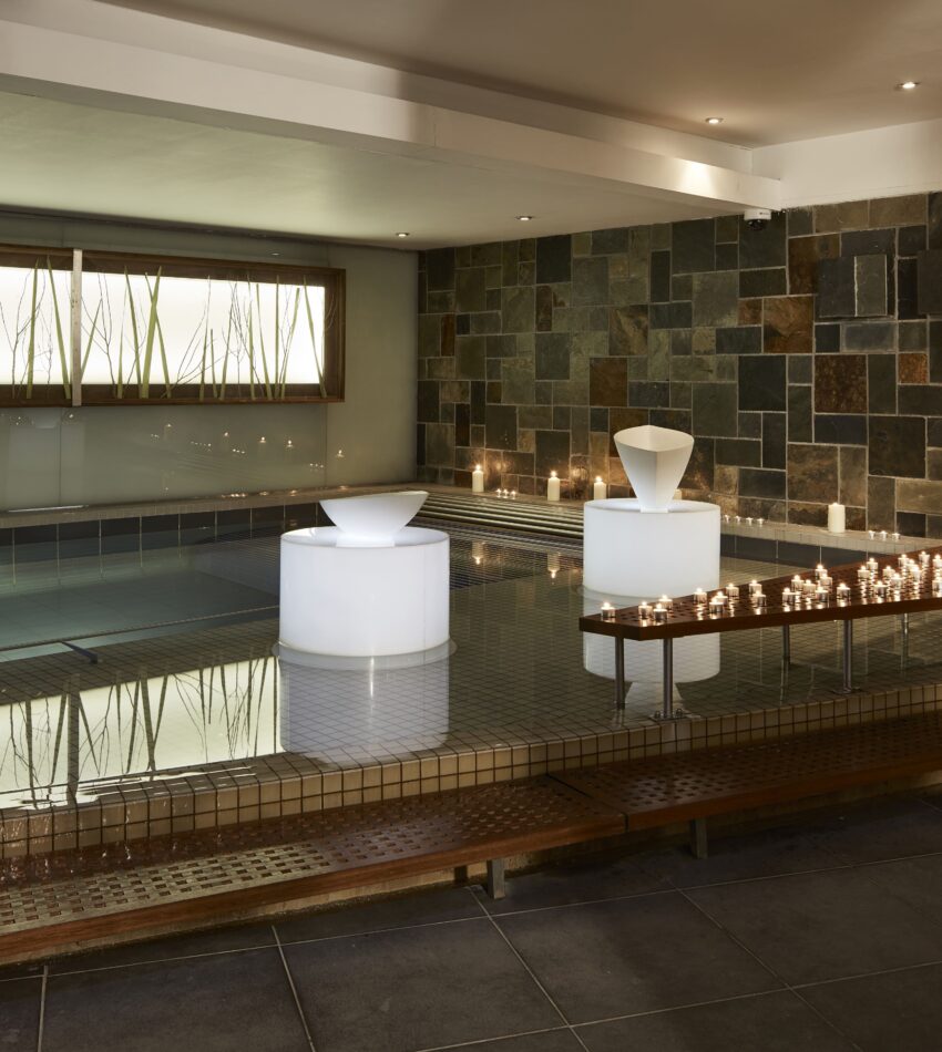 Indoor spa with tiled walls and floor, a pool, three illuminated white sculptures, candles on a wooden platform, and soft overhead lighting.