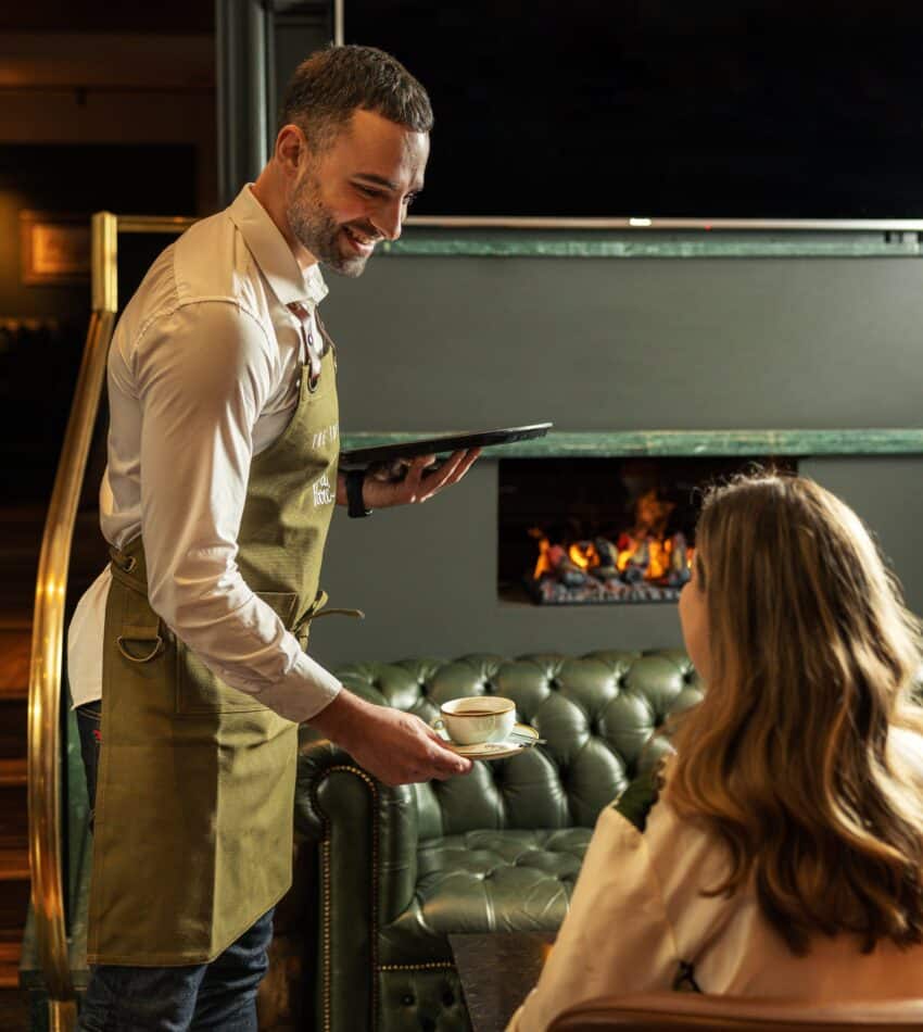 A waiter in an apron serves a cup of coffee to a seated woman in a cosy room with a green sofa and a lit fireplace.