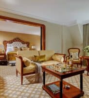 Elegant hotel suite with a sitting area, coffee table, armchairs, and a bed in the adjoining room. Large window provides natural light and a scenic view.