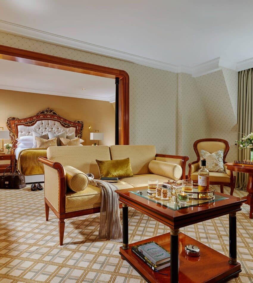 Elegant hotel suite with a sitting area, coffee table, armchairs, and a bed in the adjoining room. Large window provides natural light and a scenic view.