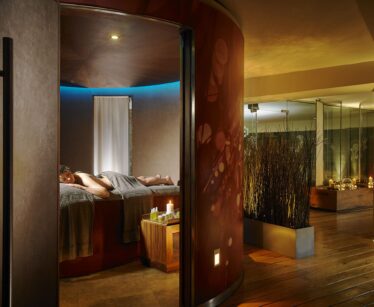 A person lies face down on a massage table in a dimly lit spa room with candles, towels, and calming décor.