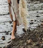 Person with bare feet standing on rocky ground beside water, wearing a beige dress and holding a long pampas grass stem.