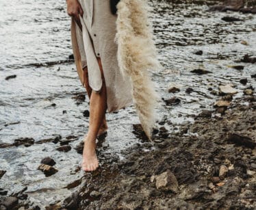 Person with bare feet standing on rocky ground beside water, wearing a beige dress and holding a long pampas grass stem.