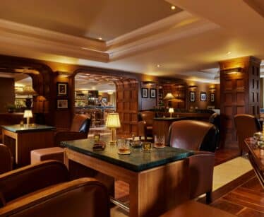 A warmly lit lounge with leather armchairs, wooden tables, and a bar in the background, featuring drinks, lamps, and wood-panelled walls.