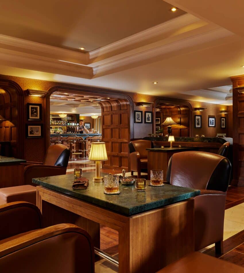 A warmly lit lounge with leather armchairs, wooden tables, and a bar in the background, featuring drinks, lamps, and wood-panelled walls.
