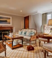 Elegant hotel suite with antique-style furniture, a coffee table set with drinks, flowers, a fireplace, and a large window allowing in natural light.