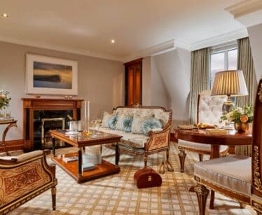 Elegant hotel suite with antique-style furniture, a coffee table set with drinks, flowers, a fireplace, and a large window allowing in natural light.