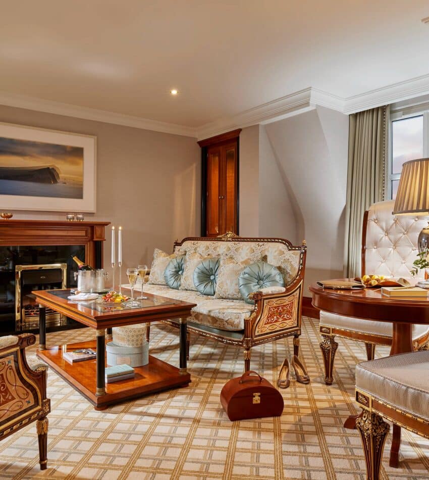 Elegant hotel suite with antique-style furniture, a coffee table set with drinks, flowers, a fireplace, and a large window allowing in natural light.