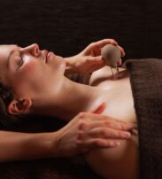 A person lying under a brown towel receives a massage, with one hand on their shoulder and another holding a round massage tool near their chest.