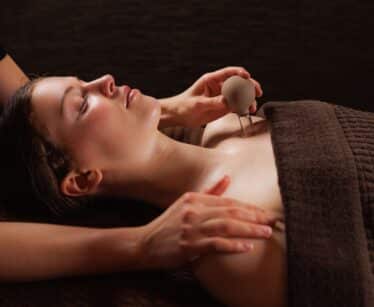 A person lying under a brown towel receives a massage, with one hand on their shoulder and another holding a round massage tool near their chest.