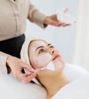 A person lies on a spa bed whilst a beautician applies a white facial mask to their face with a brush.