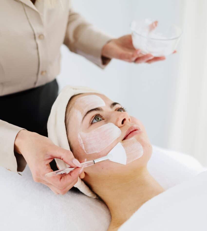 A person lying down with a white towel wrapped around their head has a face mask applied by another person using a brush.