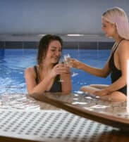 Two women in swimsuits sit in an indoor pool, smiling and clinking glasses, with water flowing from a spout and soft lighting nearby.
