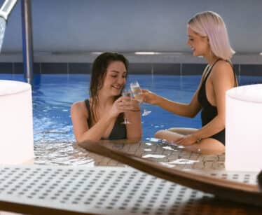 Two women in swimsuits sit in an indoor pool, smiling and clinking glasses, with water flowing from a spout and soft lighting nearby.