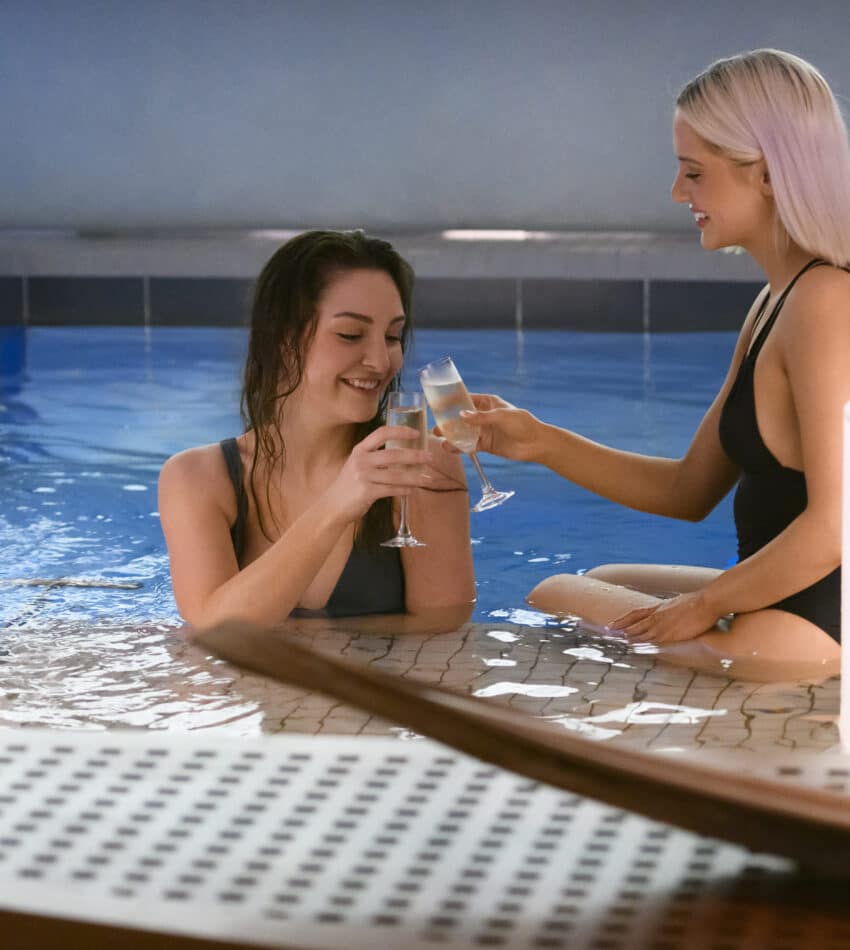 Two women in swimsuits sit in an indoor pool, smiling and clinking glasses, with water flowing from a spout and soft lighting nearby.