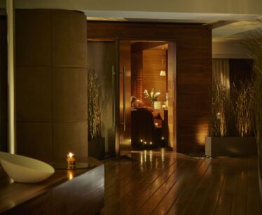 A dimly lit, modern spa lounge with wooden floors, tall plants, candles, and a person relaxing inside a warmly lit room.