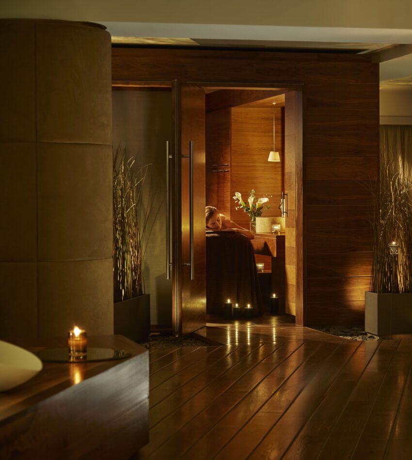 A dimly lit, modern spa lounge with wooden floors, tall plants, candles, and a person relaxing inside a warmly lit room.