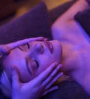 A person lying with eyes closed receives a facial massage, wrapped in a towel, under soft purple lighting.