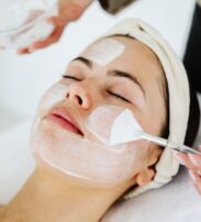 A person lies with eyes closed while a skincare professional applies a white facial mask with a brush; a towel is wrapped round the head.