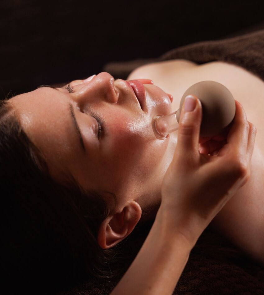 A person is lying down with eyes closed while a hand applies a glass cupping tool to their face during a facial treatment.