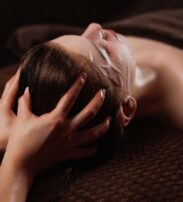 Person lying down with a facial mask, receiving a head massage, covered with a brown towel on a brown textured surface.