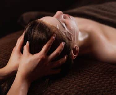 Person lying down with a facial mask, receiving a head massage, covered with a brown towel on a brown textured surface.