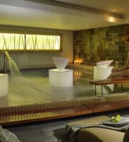 A woman in a black swimming costume sits on a bench beside an indoor spa pool, with lit candles and modern water features creating a calm atmosphere.
