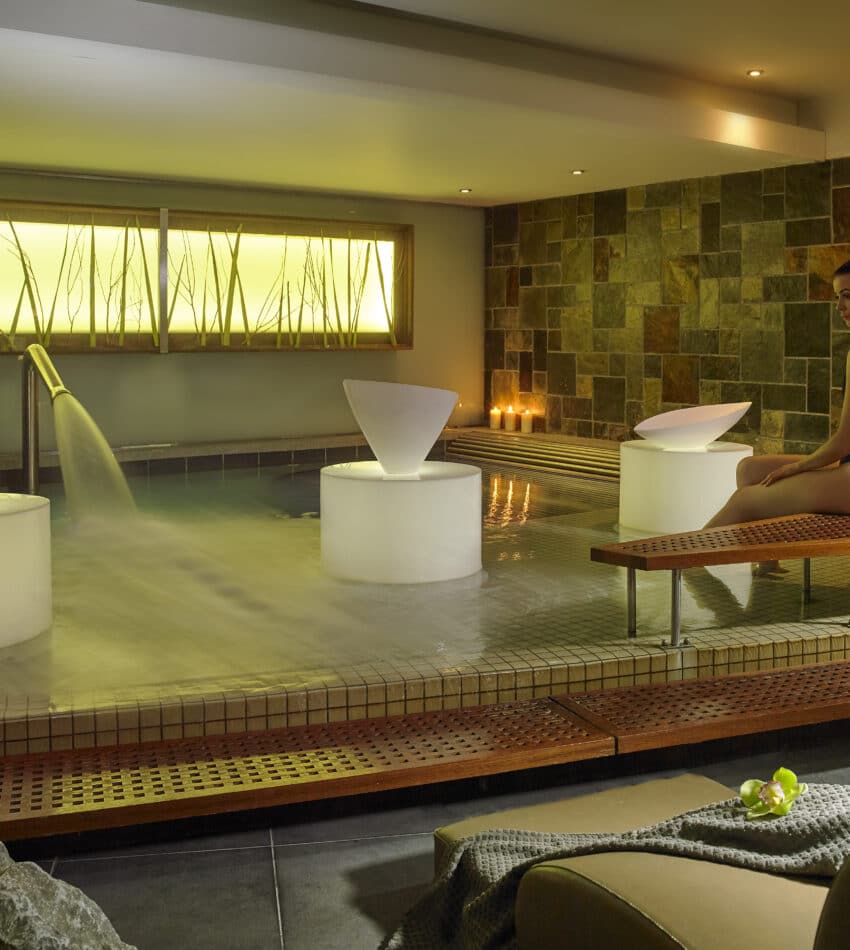 A woman in a black swimming costume sits on a bench beside an indoor spa pool, with lit candles and modern water features creating a calm atmosphere.